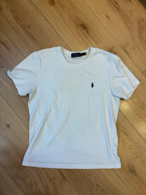 Polo Ralph Lauren T-shirt  - Tröjan är lite sönder  jag har markerat de de i bild 3.                                                Klassisk vit t-shirt från Polo Ralph Lauren med rund hals och den ikoniska svarta logotypen broderad på bröstet. Tillverkad i mjuk bomull och har en normal passform. Perfekt basplagg för alla tillfällen.  