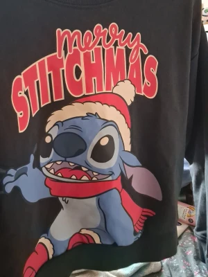 Svart Stitch julhoodie - Cool svart hoodie med stort tryck av Stitch i tomteluva och röd halsduk samt texten 'Merry Stitchmas' i rött och vitt. Perfekt för dig som älskar Disney och vill ha en julig vibe. Mjuk insida och ribbade muddar. Materialet känns som bomullsblandning.
