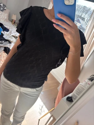 Svart topp med volangärmar - Snygg svart topp med diskret rutmönster och volangdetaljer på ärmarna. Toppen har rund halsringning och är gjord i ett lätt, något glansigt tyg som ger en elegant känsla. Perfekt att styla med jeans eller kjol för en trendig look.