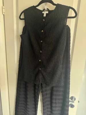 Svart set - Säljer ett svart set från med strukturerat tyg. Toppen är ärmlös med rund halsringning och knappar framtill. Byxorna är vida och har liknande material. Är man intresserad av endast ett plagg kan man skriva till mig. Vesten är i storlek S och byxorna är i storlek M.