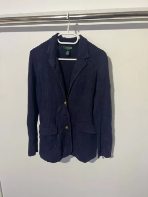 Mörkblå kavaj från Lauren Ralph Lauren - Stilren mörkblå kavaj från Lauren Ralph Lauren med två guldknappar framtill och klassisk krage. Kavajen har två fickor med lock och är tillverkad i ett strukturerat tyg som ger en snygg look. Perfekt för dig som vill ha en tidlös och elegant stil.