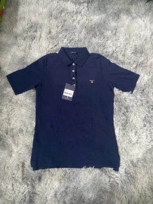 Mörkblå pikétröja från Gant - Klassisk mörkblå pikétröja från Gant med krage, korta ärmar och knappar framtill. Tröjan har en liten broderad logga på bröstet och är tillverkad i bomull för en bekväm känsla. Perfekt till jeans eller shorts.Den är helt ny med prislapp på och kostade 899kr.