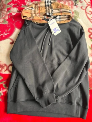 Svart hoodie från Burberry med rutig huva - Snygg svart hoodie från Burberry med den klassiska beige-rutiga huvan och dragsko. Hoodien har en avslappnad passform, ribbade muddar och en stor ficka framtill. Tillverkad i mjukt bomullsmaterial för maximal komfort. Perfekt för en trendig streetwear-look.