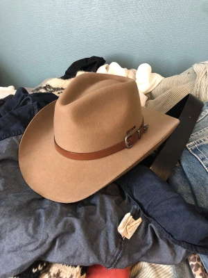 Beige cowboyhatt från Stetson - Säljer en beige cowboyhatt från Stetson i filtmaterial. Hatten har bred brätte och ett brunt läderband med snygga silverdetaljer runt kronan. Perfekt för dig som vill sticka ut med westernstil och klassisk look.