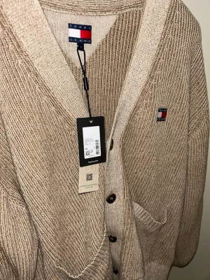 Beige kofta från Tommy Jeans - Snygg beige stickad kofta från Tommy Jeans med v-ringning, knappar. Storlek S. Passar nu till våren. 