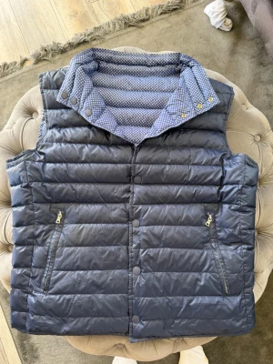 Vändbar väst från hacket london  - Snygg vändbar dunväst i blått med ena sidan enfärgad och andra sidan småprickigt mönster. Västen har hög krage, tryckknappar och dragkedjefickor. Perfekt för dig som vill variera din stil och hålla dig varm. Materialet är quiltat och lätt.