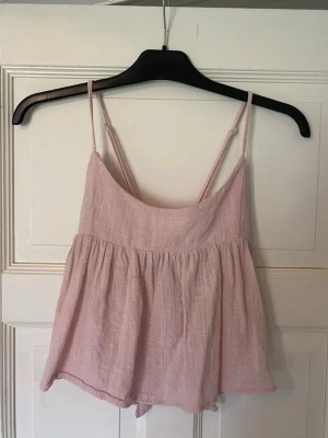 Ljusrosa linnetopp med knyt i ryggen - Supersöt ljusrosa linnetopp med tunna axelband och en snygg knytning i ryggen. Toppen har en luftig och flowy passform, perfekt för varma dagar. Tillverkad i ett mjukt och lätt material som känns skönt mot huden.
