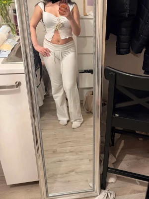 Vit croppad snörtopp med rosett - Supersnygg vit croppad topp med snörning framtill och en söt rosett. Toppen har breda axelband och en v-ringad urringning som ger en feminin look. Perfekt att matcha med höga byxor eller kjol för en trendig outfit.