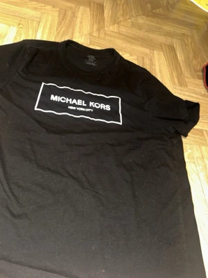 Svart Michael Kors t-shirt - Snygg svart t-shirt från Michael Kors med vit logga och texten 'NEW YORK CITY' på bröstet. Klassisk rund halsringning och korta ärmar. Tillverkad i mjuk bomull som känns skön mot huden. Perfekt för dig som gillar stilrena och trendiga plagg. tshirten är köpt i usa för ett par år sedan då jag fick som födelsedagspresent men hsr nu växt ur skicket är hyfsat bra ändå för hur länge jag haft den priset kan diskuteras. frakt eller upphämtning spelar ingen roll för mig.