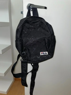 Svart ryggsäck från FILA - Snygg svart ryggsäck från FILA med diskret mönster och logga framtill. Väskan har justerbara axelband med FILA-tryck, ett stort huvudfack och en mindre ficka framtill med dragkedja. Perfekt storlek för vardagens essentials och enkel att matcha.