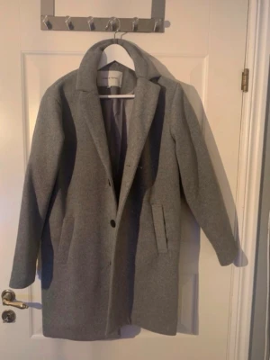 Grå klassisk ullkappa Selected Femme - Stilren grå kappa från Studio Total i ullblandning. Kappan har klassisk krage, två fickor framtill och stängs med knapp. Rak passform och helfodrad för extra komfort. Perfekt för kyliga dagar och enkel att matcha med olika outfits. Aldrig använd någonsin bara hängt i garderoben. Storlek 158/164