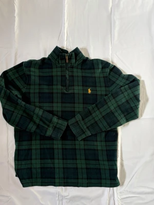 Grönrutig halvzip från Ralph Lauren - Mörkgrön och svartrutig tröja från Ralph Lauren med halv dragkedja och klassisk gul broderad logga på bröstet. Tröjan har hög krage och långa ärmar, perfekt för lager på lager. Tidlös design som funkar till det mesta.