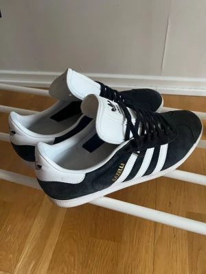 Adidas Gazelle svarta sneakers - Klassiska Adidas Gazelle sneakers i svart mocka med vita detaljer och de ikoniska tre ränderna på sidan. Låg modell med vit sula och vit hälflik. Guldtexten 'GAZELLE' på sidan ger extra stilpoäng. Perfekta för en clean look.
