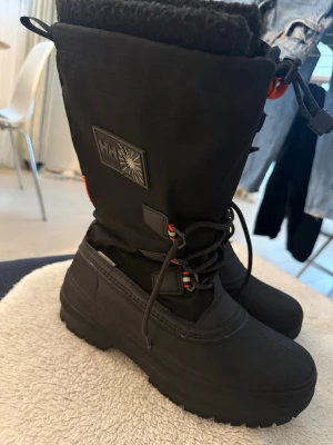 Svarta moon boots från Helly Hansen - Säljer ett par svarta moon boots från Helly Hansen med grov sula och snörning framtill. Bootsens ovandel är i slitstark textil och nederdelen i gummi, perfekta för kalla dagar. Stora HH-loggor på sidan och sulan ger en sportig vibe.