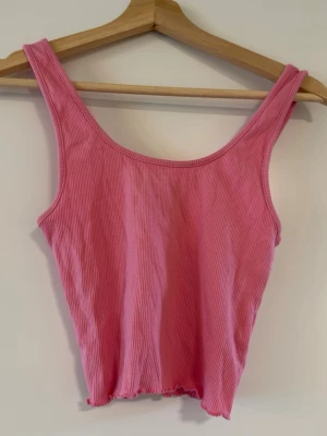 Rosa ribbad linnetopp - Säljer en söt rosa ribbad linnetopp med croppad passform. Toppen är ärmlös med breda axelband och har vågig kant nertill för en extra detalj. Perfekt att styla till jeans eller kjol för en chill look.