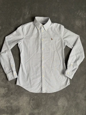 Randig skjorta från Ralph Lauren - Klassisk långärmad skjorta från Ralph Lauren med tunna blå och vita ränder. Skjortan har button-down krage, vita knappar och den ikoniska broderade loggan på bröstet. Tillverkad i mjuk bomull som känns skön mot huden. Är i helt nyskick, har bara använts några gånger! Perfekt för en clean och stilren look till sommaren!     Storlek S. Måtten: längd 60cm, bredd 45cm.       Hör av dig vid minsta fundering!😊 