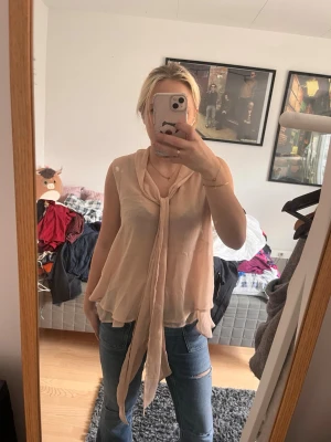 Ljusbeige ärmlös blus med knytband - Säljer en ljusbeige, ärmlös blus i ett tunt och luftigt material. Blusen har ett långt knytband vid halsen som ger en snygg detalj och ett avslappnat fall. Perfekt att styla med jeans eller kjol för en trendig look.
