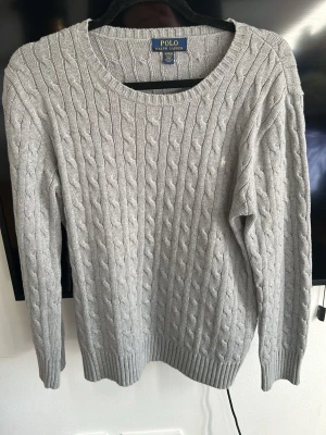 Grå kabelstickad tröja Polo Ralph Lauren - Säljer en grå kabelstickad tröja från Polo Ralph Lauren. Tröjan har rund halsringning, långa ärmar och ribbade muddar vid ärmslut och nederkant. Klassisk och stilren design med diskret broderad logga på bröstet. Perfekt till jeans eller chinos.