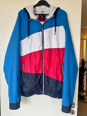 Vindjacka - Snygg vindjacka från Pull&Bear med blå, vit, röd och mörkblå färgblock. Jackan har huva, dragkedja framtill och två fickor med dragkedja. Materialet är lätt och luftig. Storlek XL. plastdelen man håller för att använda kedjan har gått halvt av men fungerar normalt (se bild). 