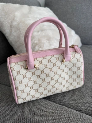 Handväska rosa/vit - Snygg handväska med Triomphe-mönster i beige på vit bakgrund och ljusrosa detaljer. Väskan har två handtag, dragkedja upptill och avtagbar axelrem. 