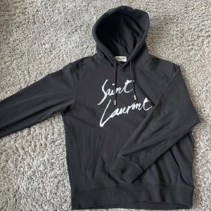 Svart hoodie från Saint Laurent - Säljer en svart hoodie från Saint Laurent med vit logga framtill. Tröjan passet runt 175-180 tröjan är i nyskick och inga defekter. De stor även saint Laurent på metal biten på snörena. Byten är väldigt intressant!!!