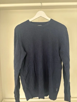 Mörkblå stickad tröja  - En enkel och stilren mörkblå stickad tröja från Jack and Jones med rund halsringning och långa ärmar. Tröjan har ribbade muddar vid ärmslut och nederkant. Perfekt att matcha med jeans eller chinos för en clean look.