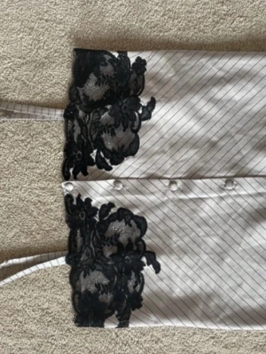 Randigt nattlinne med spetsdetaljer - Snyggt nattlinne med tunna axelband i vitt tyg med tunna svarta ränder. Dekorerat med svart blommig spets upptill och nedtill. Knappar framtill ger en extra touch. Perfekt för en lyxig och bekväm känsla på kvällen.