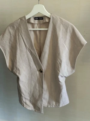 Beige väst med v-ringning från - Stilren beige väst  med v-ringning och en knapp framtill. Västen har en lös passform och är tillverkad i ett lätt material som ger en avslappnad look. Perfekt att styla över en t-shirt eller skjorta för en trendig vibe.