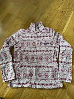 Patagonia, dam, L mönstrad fleecejacka  - Mysig fleecejacka från Patagonia i vitt och rosa med ett snyggt blommigt mönster. Jackan har dragkedja. Perfekt för kyliga dagar och enkel att slänga på sig över en hoodie eller t-shirt.