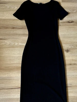 Svart ribbad klänning från Dasy  - Svart ribbad klänning från Dasy med korta ärmar och fyrkantig ringning. Klänningen är figurnära och har en enkel, stilren design som passar perfekt till många olika tillfällen. Materialet är stretchigt och bekvämt.