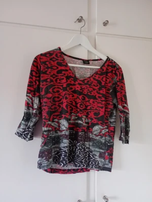 Mönstrad topp från Chic Celebes - Unik topp från Chic Celebes med röd och svart abstrakt mönster, v-ringad hals och trekvartsärm. Materialet känns mjukt och stretchigt, perfekt för dig som gillar färgstarka plagg med cool design.