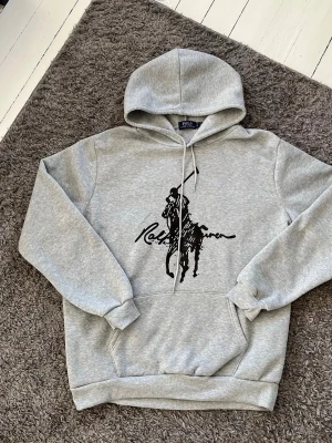 Grå hoodie från Polo Ralph Lauren - Snygg grå hoodie från Polo Ralph Lauren med stor svart polospelare och logga på bröstet. Klassisk känguruficka framtill, ribbade muddar och justerbar dragsko i huvan. Perfekt för en avslappnad och trendig stil.