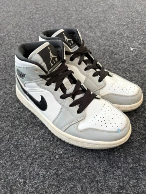 Nike Air Jordan 1 Mid grå/vit - Snygga Nike Air Jordan 1 Mid sneakers i vitt och ljusgrått läder med svarta detaljer och svart Swoosh. Klassisk Jordan-logga på sidan och svartvita skosnören. Perfekta för dig som vill ha en ikonisk och stilren look.