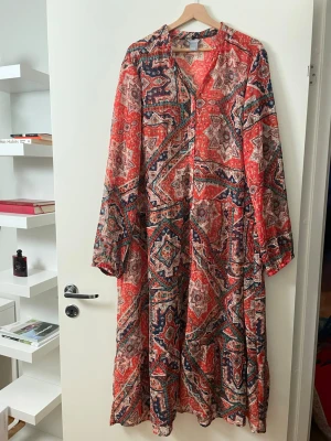 Lindex Mönstrad långklänning i bomull - Färgstark långklänning med orientaliskt inspirerat mönster i rött, blått och beige. Klänningen har lång ärm, v-ringning och knappar framtill. Tillverkad i tunn, luftig bomull som passar perfekt för varma dagar. Storlek är XXL men kan även passa för de som har M, L, XL
