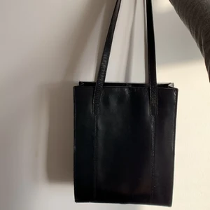 Håndtaske i sort læder  - Tasken har lange stropper, har plads til en 13’’ bærbar. Lukkes sammen med en magnetknap foroven. 