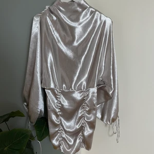 Silvrig klänning från ASOS Design - Silvrig klänning med öppen rygg/ballongärmar som man knyter. Två knappar runt halsen för att stänga samt dragkedja i midjan. Då detta är min egna bilder kan man få fler utan den på. Storlek XS. Bortse från guldiga bandet runt, hör ej till klänningen. 