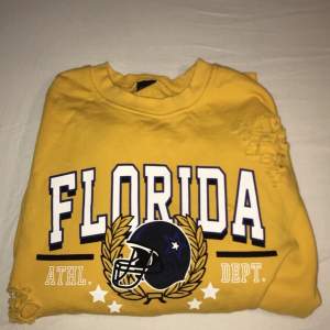 En gul långärmad tröja med trycket ”Florida” tvärs över bröstet. Det är ”slitningar” över hela tröjan samt två vita sträck på båda ärmarna. Storleken är 36/S. 