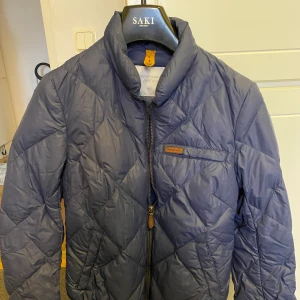 Peak dunjacka strl XS - Dunjacka från Peak performance i fint skick. Mörkblå med brun läderdetalj på bröstet med märket samt skinnflärp på dragkedjan. Perfekt vår samt höst jacka.