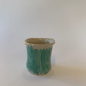 Handgjord kopp med grönt fält - En handgjord kopp i keramik med en naturlig sandfärg och ett grönt fält. Denna kopp saknar öra men passar dock bra i handen. 