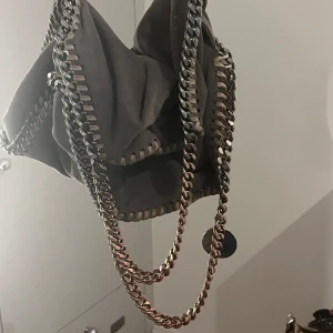 Stella McCartney väska  - Äkta Stella McCartney falabella väska i grå och silver. De korta Banden är rostiga men går att fixa med rostfärg. Litet hål se bild. Finns äkthetskod i väskan. Använd väska säljs i befintligt skick. Inga byten. Det långa bandet är lite ner kortad 