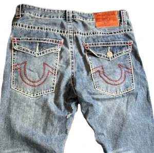 True Religion jeans - Blå true religion jeans med röda detaljer i bra skick. Mått: ytterbenlängd 112cm, midjelångd 48cm, innerbenlängd 82cm.  Defect: Vänsterbakficka saknar knapp. Litet hål på vänster innersida byxben.