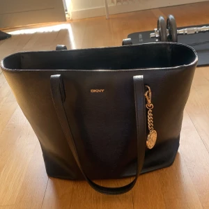 DKNY, Donna Karan,  Handväska, nyskick.  - Säljer min DKNY, Donna Karan, väska i mycket gott skick. Svart färg. Det finns även två mindre fack i väskan en med  dragkedja.  Logo hänget ni ser på väskan kan man ta bort om man vill. Stort utrymme i väskan. OBS! Nypris 2500kr. 