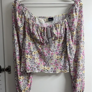 Helt ny blommig blus - En söt blus med blommigt mönster och en fin detalj med snörning i fram. Blusen har långa ärmar och en liten volang. Helt oanvänd och lappar sitter kvar