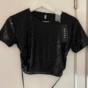 Paljettopp med knytning i ryggen - Paljett-topp från Motel. Öppen i ryggen som man knyter. Aldrig använd. Kapparna är kvar. 