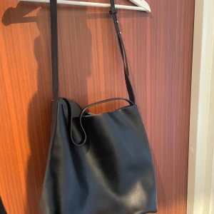 Svart läderväska  - Svart läderväska från NA-KD. Hade den början vaxade kanter upptill samt runt bandet, men detta började spricka så jag plockade bort det för en mjukare stil och följsamhet i väskan. Är kvar på det kortare bandet. Nypris 899 kr. 