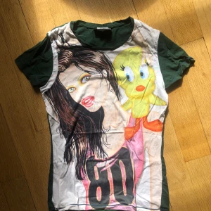 Grön t-shirt med tryck från Étoile du Monde - En grön t-shirt med tryck från Étoile du Monde. Framsidan har en färgglad illustration av en kvinna och en tecknad figur, samt siffran 80. Baksidan är enfärgad grön med vita detaljer på ärmarna.