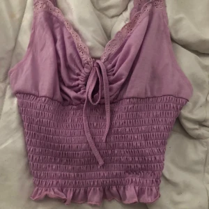 Lila topp med spetsdetaljer - En lila topp med spetsdetaljer och knytning framtill. Toppen har en smockad nederdel och volangkant.