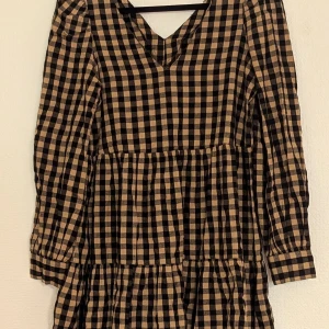 Rutig klänning - En rutig klänning med svart och beige mönster. Klänningen har långa ärmar och en v-ringad hals. Den är av normal passform och har en avslappnad stil. hemmasydd: o= 94cm