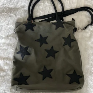 Väska - Rymlig väska, med flera fickor på insidan och ett på baksidan. Har band i läder, både ett längre axelband och ett kortare som funkar både för att hålla väskan i armvecket och på axeln. Stjärnor bara på framsidan.