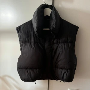 Svart dunväst - En svart dunväst med hög krage och dragkedja. Västen är vadderad och ser varm och bekväm ut. Den är i en kortare modell och storlek L men passar egemtligen S-M så skriver de i storleken